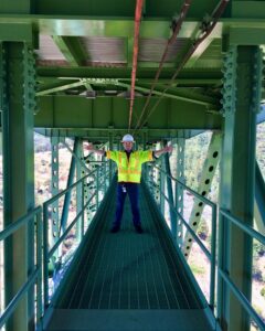 A man in a yellow vest with his arms extended is standing under the frame of a bridge